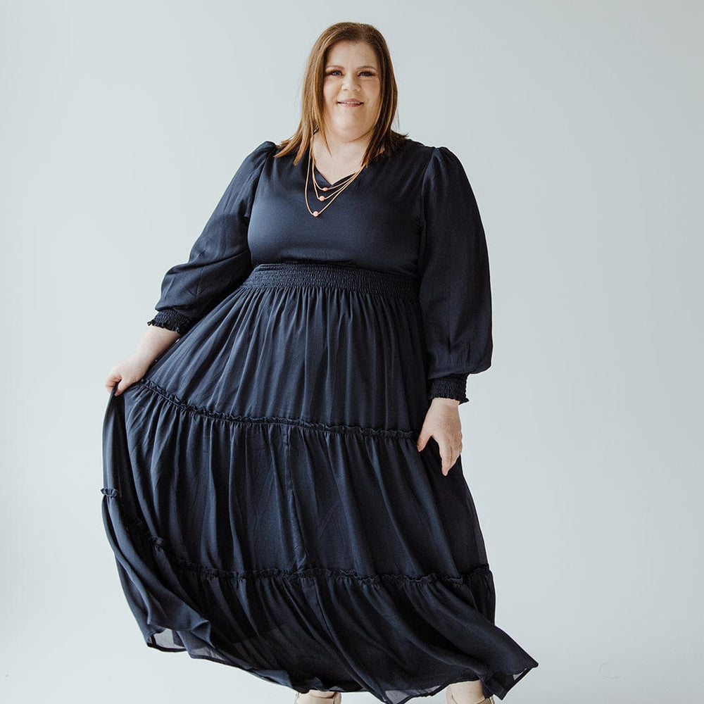 A person wearing a long, flowing black V-neck dress with a tiered skirt and beige sandals poses against a plain background.