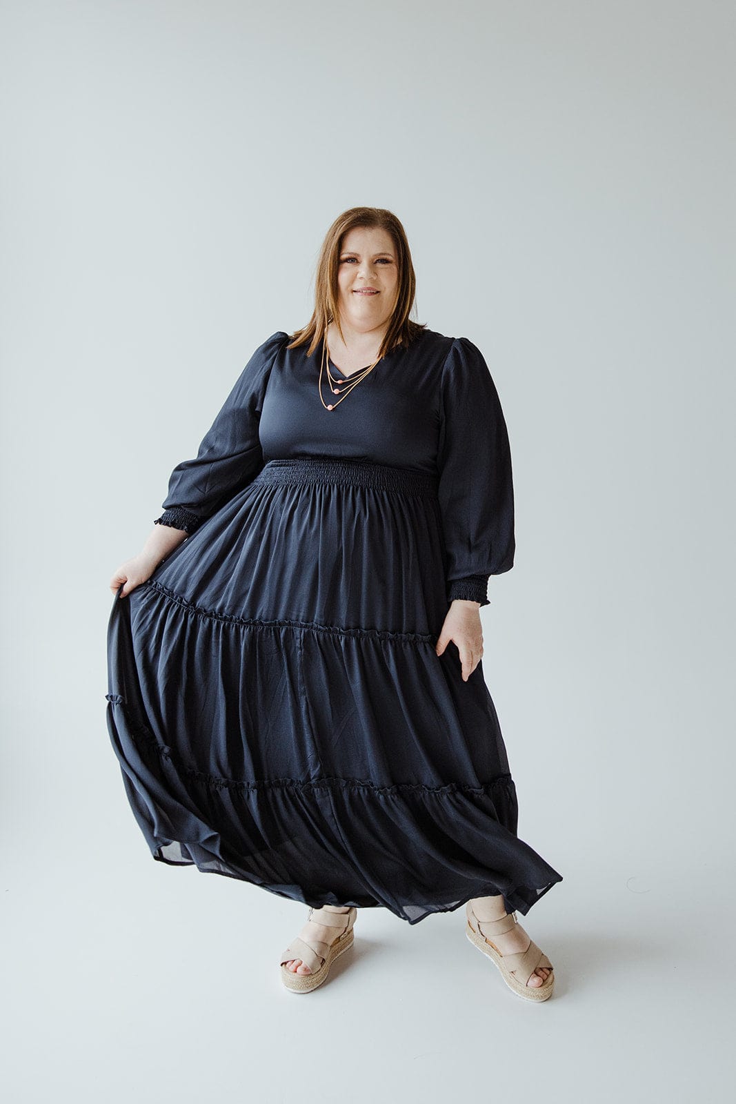 A person wearing a long, flowing black V-neck dress with a tiered skirt and beige sandals poses against a plain background.