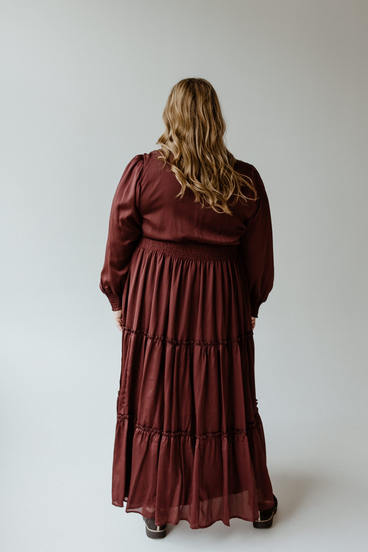 A person with long hair wearing a Zinfandel blouson dress featuring a smocked waist stands facing away, showcasing the elegant flow of the maroon fabric.