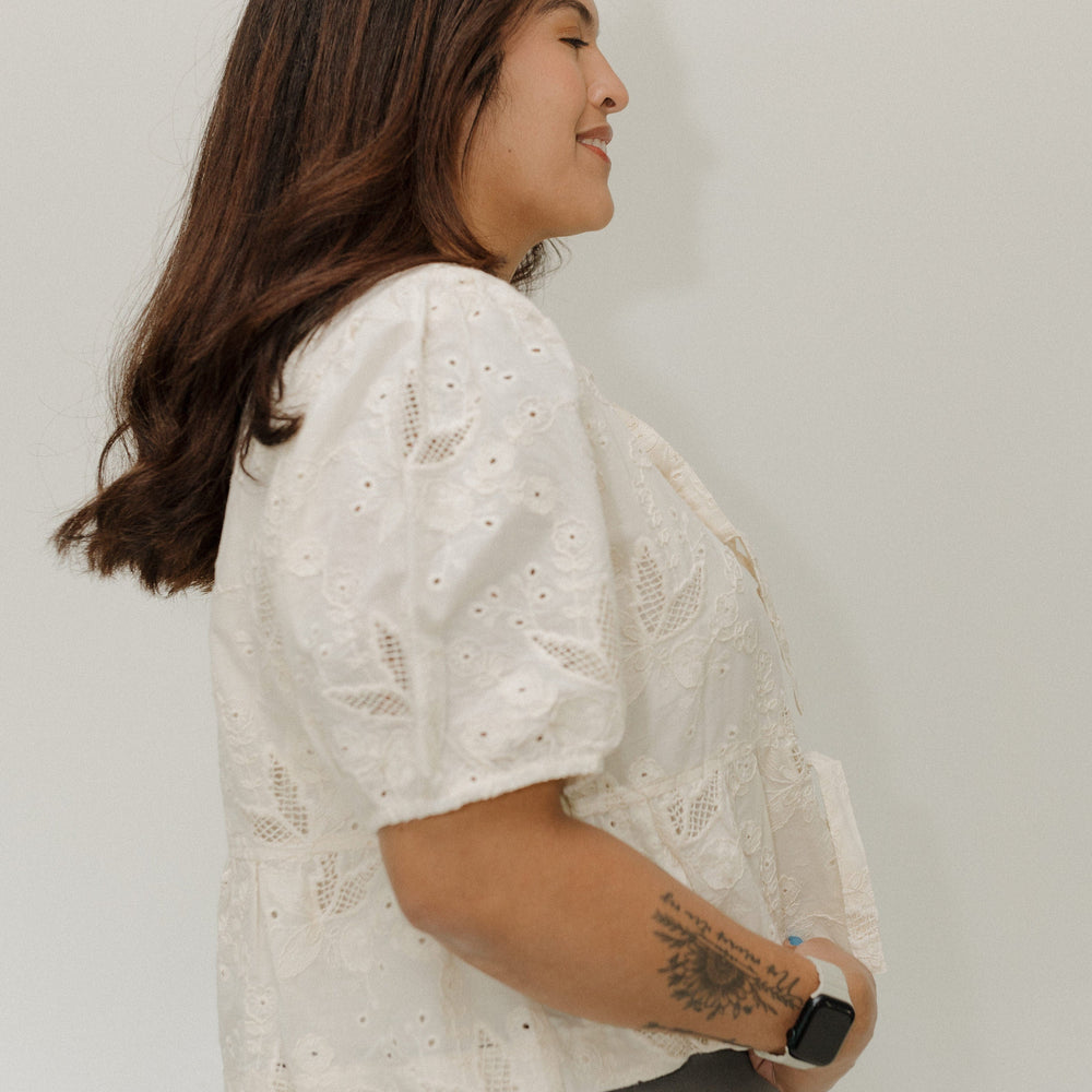 Woman wearing a white lace blouse and gray pants against a plain background