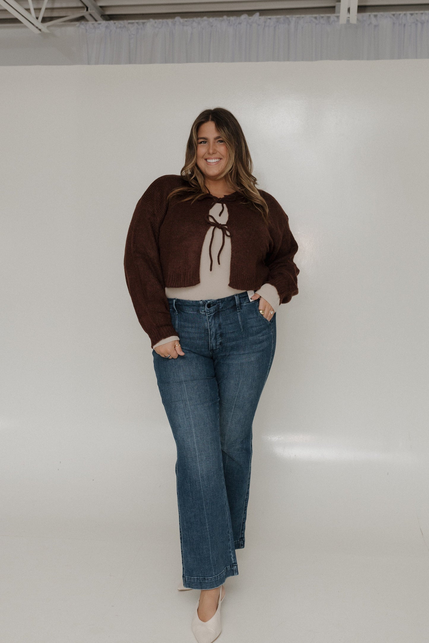 Woman wearing a brown sweater and blue jeans standing against a white wall