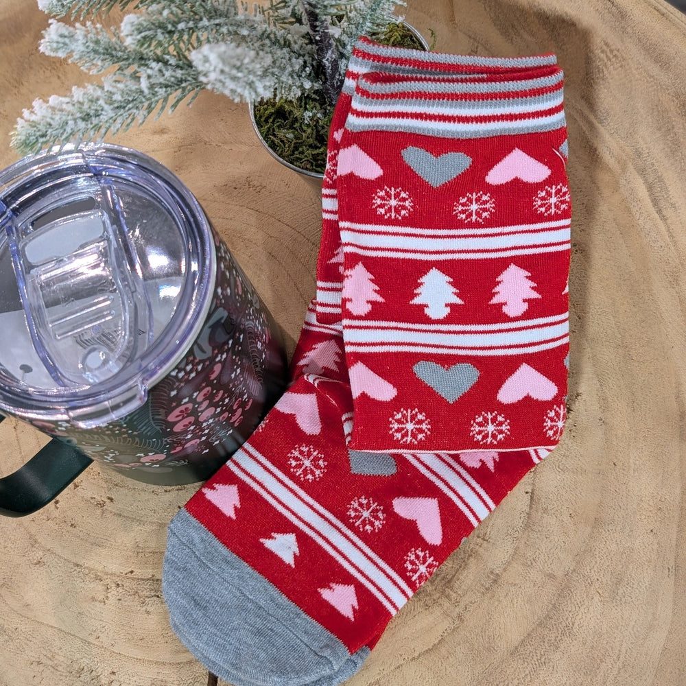 Red holiday-themed socks with hearts, snowflakes, and tree patterns are placed on a wooden surface next to a clear-lidded travel mug. Festive colors come alive as a small frosted evergreen branch decorates the cozy scene.