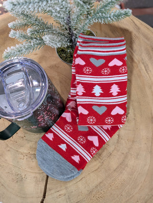 Red holiday-themed socks with hearts, snowflakes, and tree patterns are placed on a wooden surface next to a clear-lidded travel mug. Festive colors come alive as a small frosted evergreen branch decorates the cozy scene.