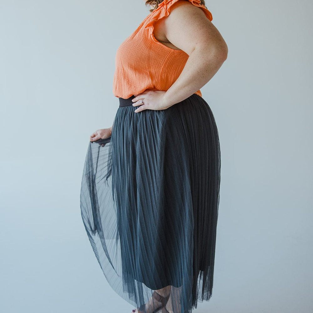 A person stands in profile wearing an orange sleeveless top, a chic Zenana TULLE MIDI SKIRT IN CHARCOAL GREY, and white sandals.