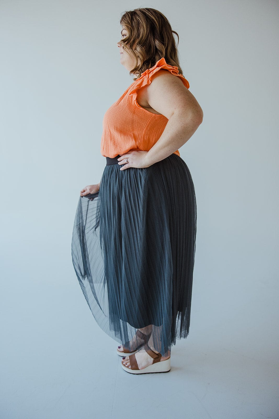 A person stands in profile wearing an orange sleeveless top, a chic Zenana TULLE MIDI SKIRT IN CHARCOAL GREY, and white sandals.