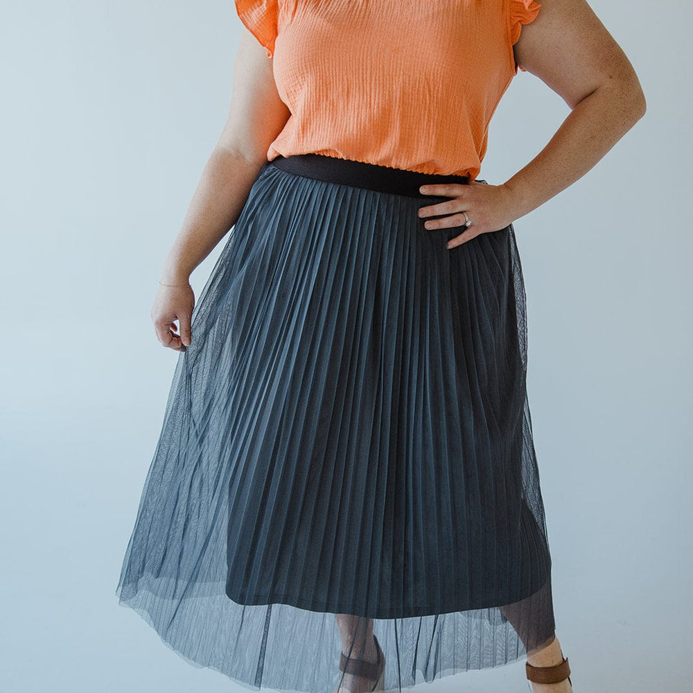 A person is wearing an orange top with ruffled sleeves, a chic TULLE MIDI SKIRT IN CHARCOAL GREY by Zenana with an elastic waist, and brown sandals. The background is white.