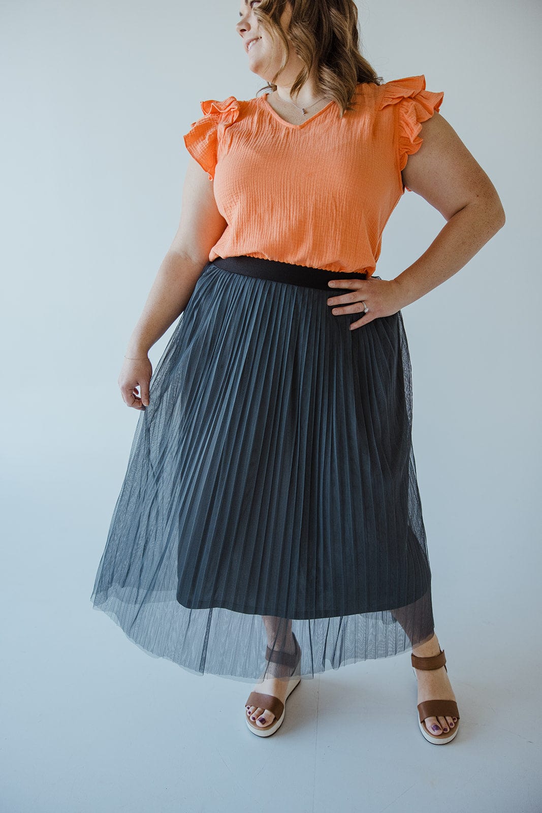 A person is wearing an orange top with ruffled sleeves, a chic TULLE MIDI SKIRT IN CHARCOAL GREY by Zenana with an elastic waist, and brown sandals. The background is white.