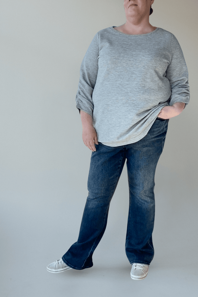 A person wearing a gray long-sleeve shirt paired with JUDY BLUE TUMMY CONTROL EMBROIDERED POCKET BOOTCUT JEANS IN TALL by Judy Blue stands against a plain backdrop, showcasing the jeans' embroidered pockets with hands casually tucked in.