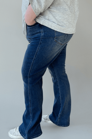 A person is wearing the Judy Blue Tummy Control Embroidered Pocket Bootcut Jeans in Tall and a gray top, with hands casually resting in the embroidered pockets, while standing sideways.