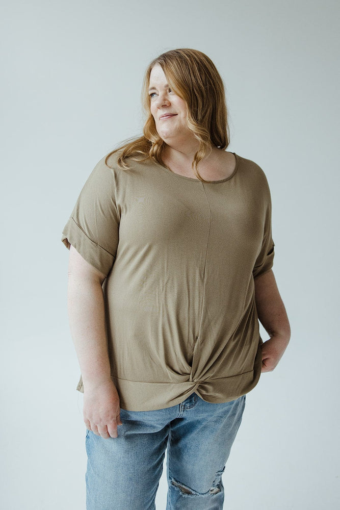 A person with long hair wearing a Zenana TWIST FRONT TEE IN SOOTHING SAGE and ripped blue jeans stands against a plain background.