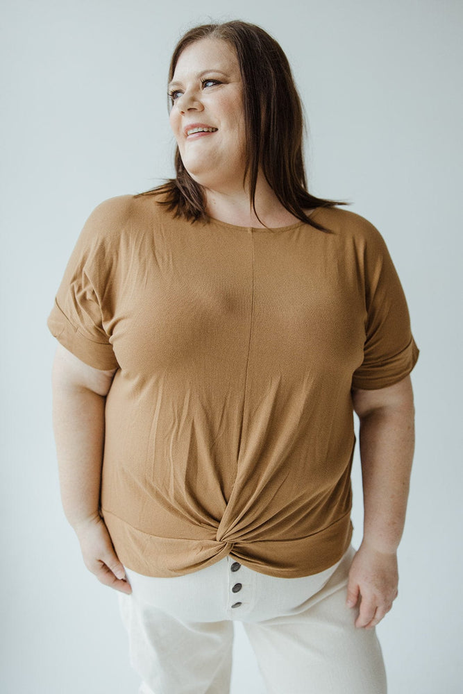 Person standing, wearing a trendy scoop neck TWIST FRONT TEE IN URBAN BRONZE by Zenana and white pants. They are smiling and looking to the side, embracing their style as a mid to plus-size fashionista.