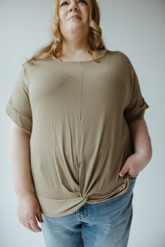 A person with long, wavy hair wearing a Zenana Twist Front Tee in Soothing Sage and blue jeans stands against a plain background.