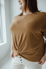 A mid to plus-size fashionista with shoulder-length brown hair stands with their back to the camera, wearing a trendy Zenana Twist Front Tee in Urban Bronze and light-colored pants against a plain background.