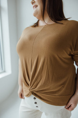 A mid to plus-size fashionista wearing a trendy scoop neck TWIST FRONT TEE IN URBAN BRONZE by Zenana and white pants stands near a window, smiling slightly.