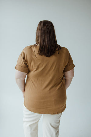 A mid to plus-size fashionista with shoulder-length brown hair stands with their back to the camera, wearing a trendy Zenana Twist Front Tee in Urban Bronze and light-colored pants against a plain background.