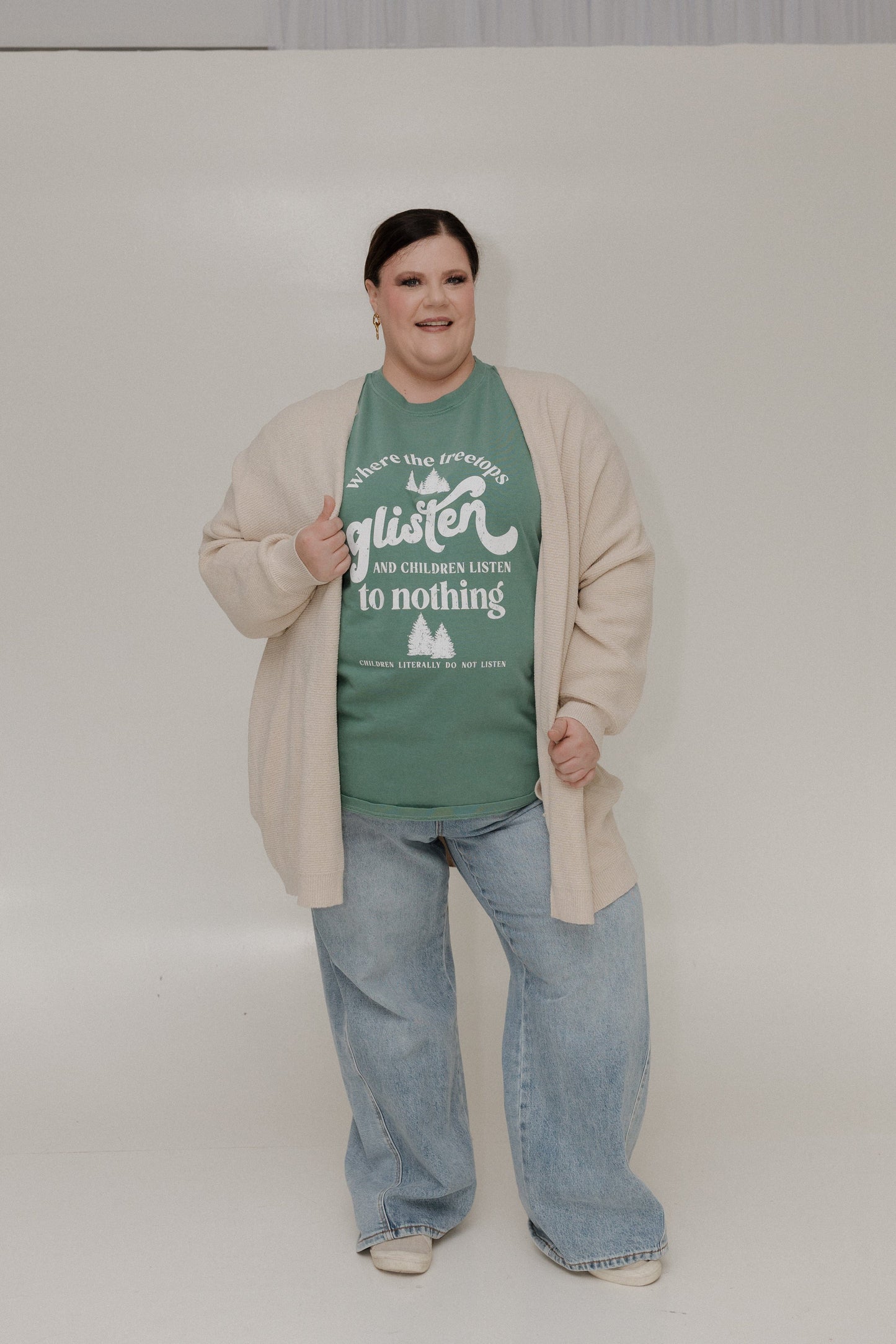 Person wearing a green t-shirt with text and a beige cardigan on a plain background