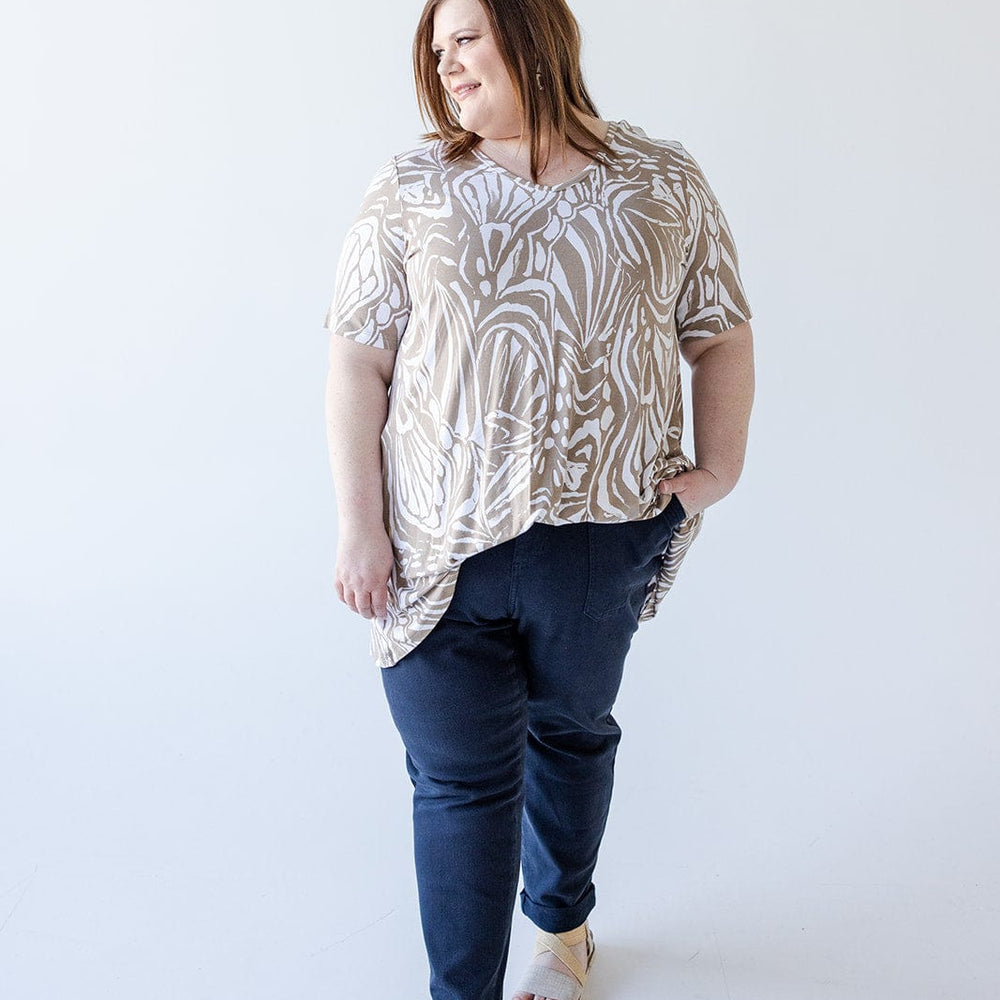 A person with long hair wearing a Love Marlow V-NECK ABSTRACT PRINT TUNIC IN SAND AND OFF-WHITE, dark pants, and sandals stands against a plain white background.