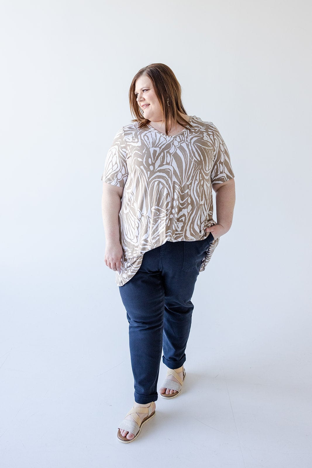 A person with long hair wearing a Love Marlow V-NECK ABSTRACT PRINT TUNIC IN SAND AND OFF-WHITE, dark pants, and sandals stands against a plain white background.