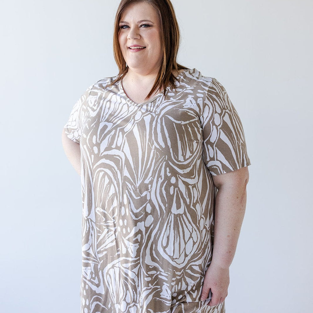 A person with shoulder-length hair is wearing a Love Marlow V-NECK ABSTRACT PRINT TUNIC IN SAND AND OFF-WHITE and dark pants, smiling against a plain white background.