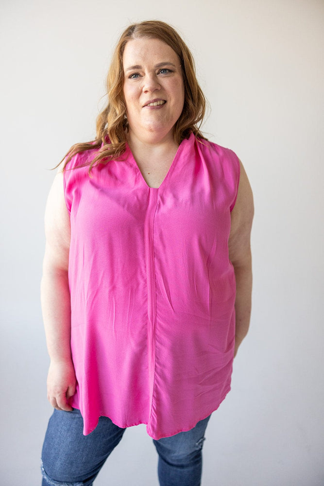 A person with long hair wearing a sleeveless pink Love Marlow V-NECK FLOWY TANK BLOUSE WITH RUCHED SHOULDER IN FRESH FUCHSIA and blue jeans stands against a plain background, showcasing summer fashion.