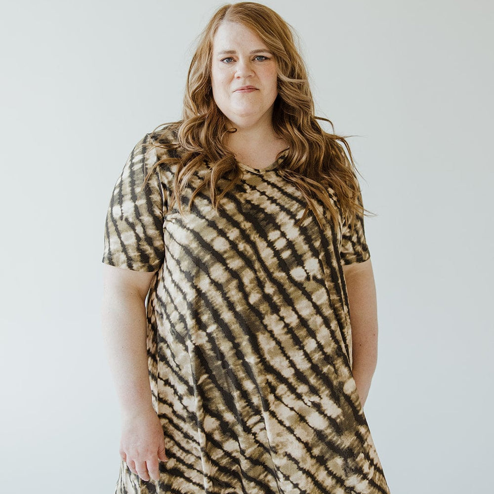 A person with long wavy hair is wearing a short-sleeved Love Marlow V-Neck Tunic Tee in Safari Grass paired with brown pants, standing against a plain background.