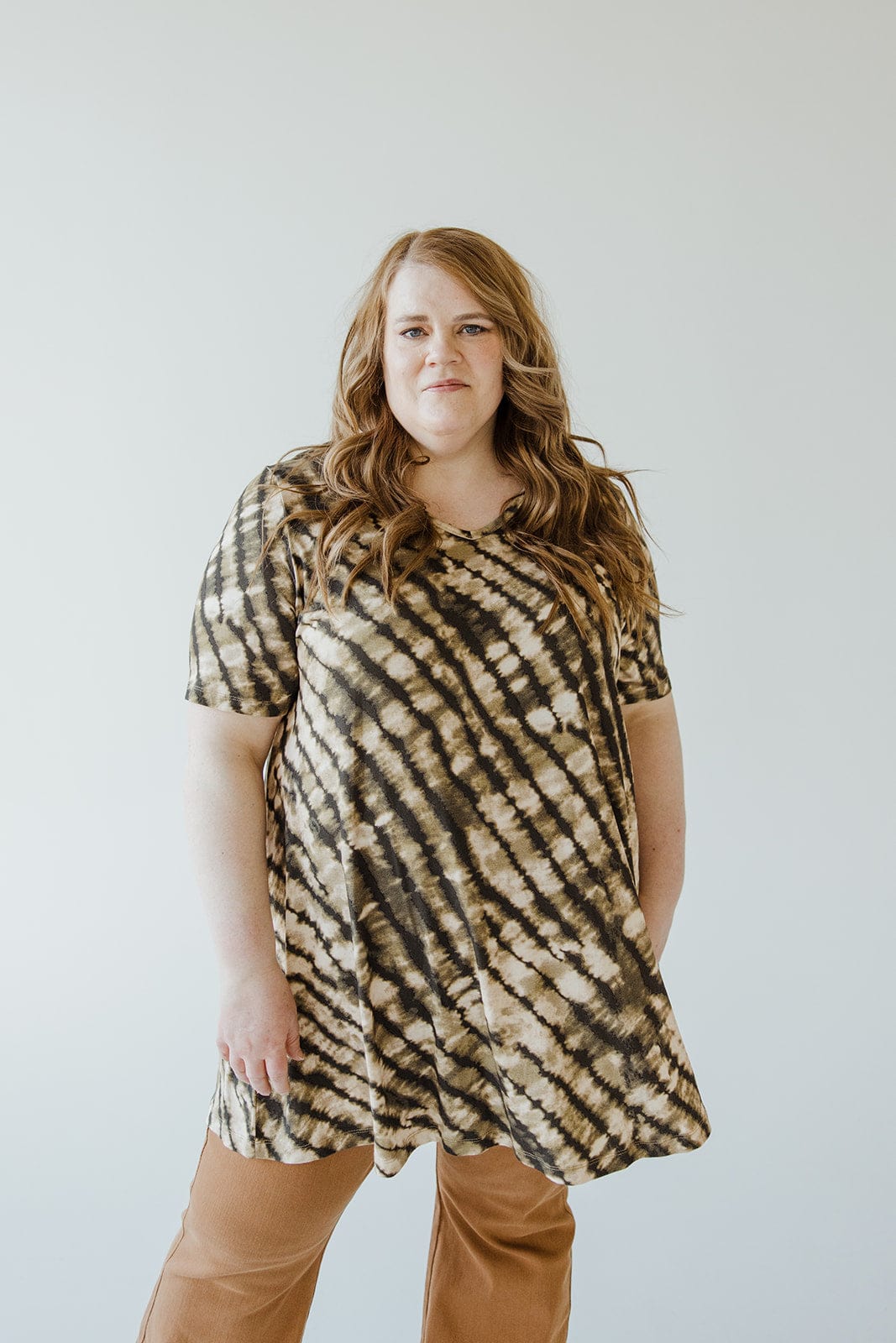 A person with long wavy hair is wearing a short-sleeved Love Marlow V-Neck Tunic Tee in Safari Grass paired with brown pants, standing against a plain background.