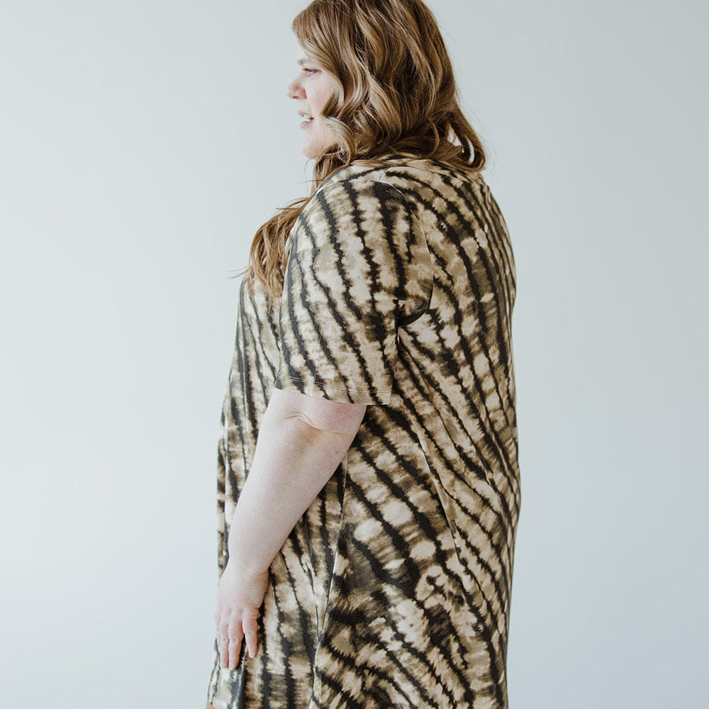 A person with long hair stands sideways wearing the V-Neck Tunic Tee in Safari Grass by Love Marlow and brown pants against a plain background, showcasing a versatile wardrobe.