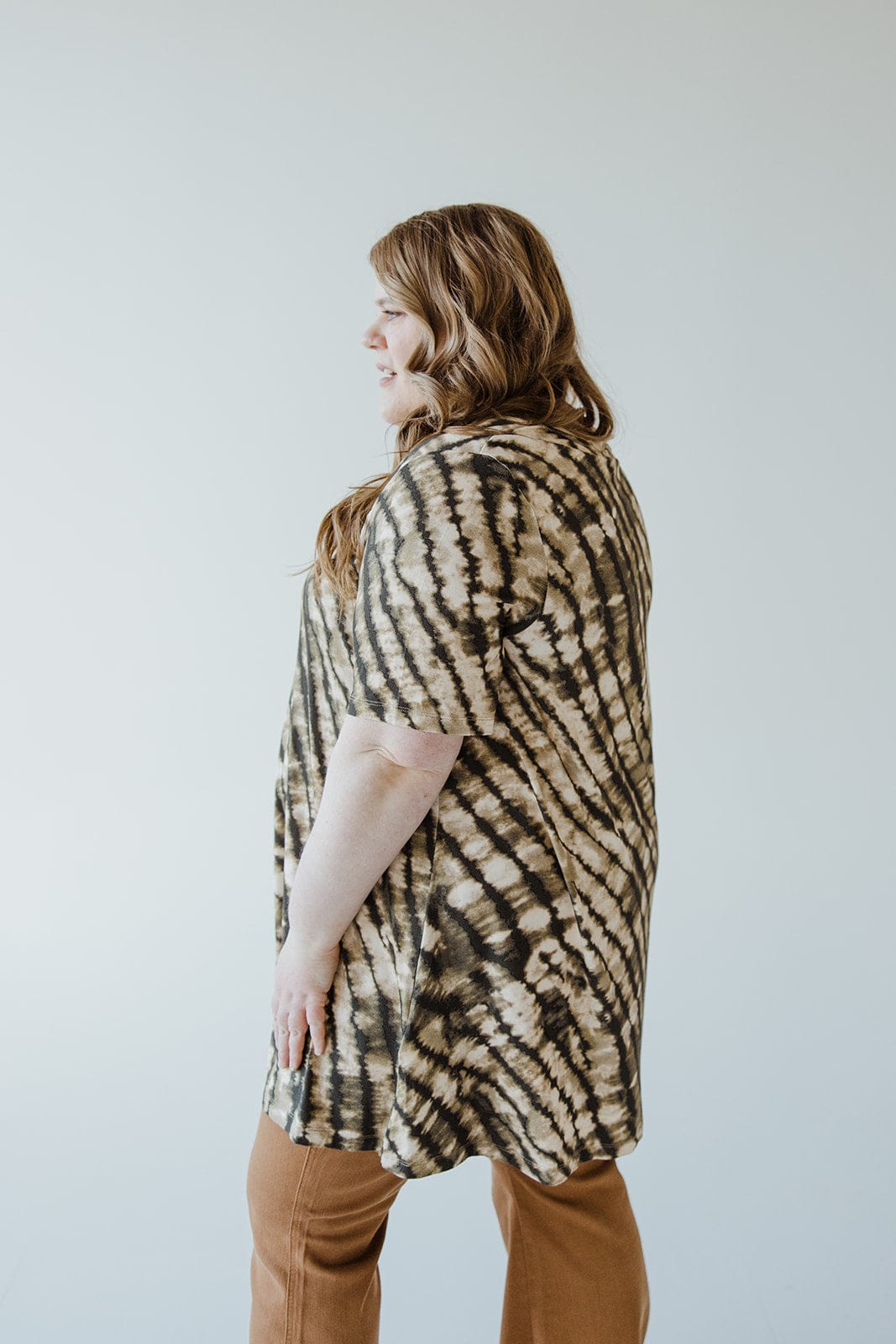 A person with long hair stands sideways wearing the V-Neck Tunic Tee in Safari Grass by Love Marlow and brown pants against a plain background, showcasing a versatile wardrobe.