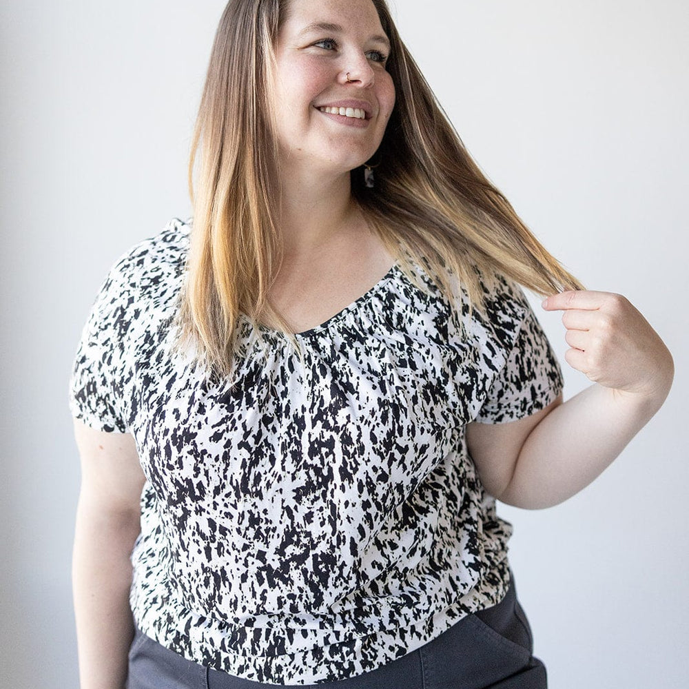 A person with long hair smiles and holds a strand of their hair. They are wearing a V-NECK TUNIC WITH ABSTRACT ANIMAL PRINT DETAIL by Love Marlow, paired perfectly with dark pants.