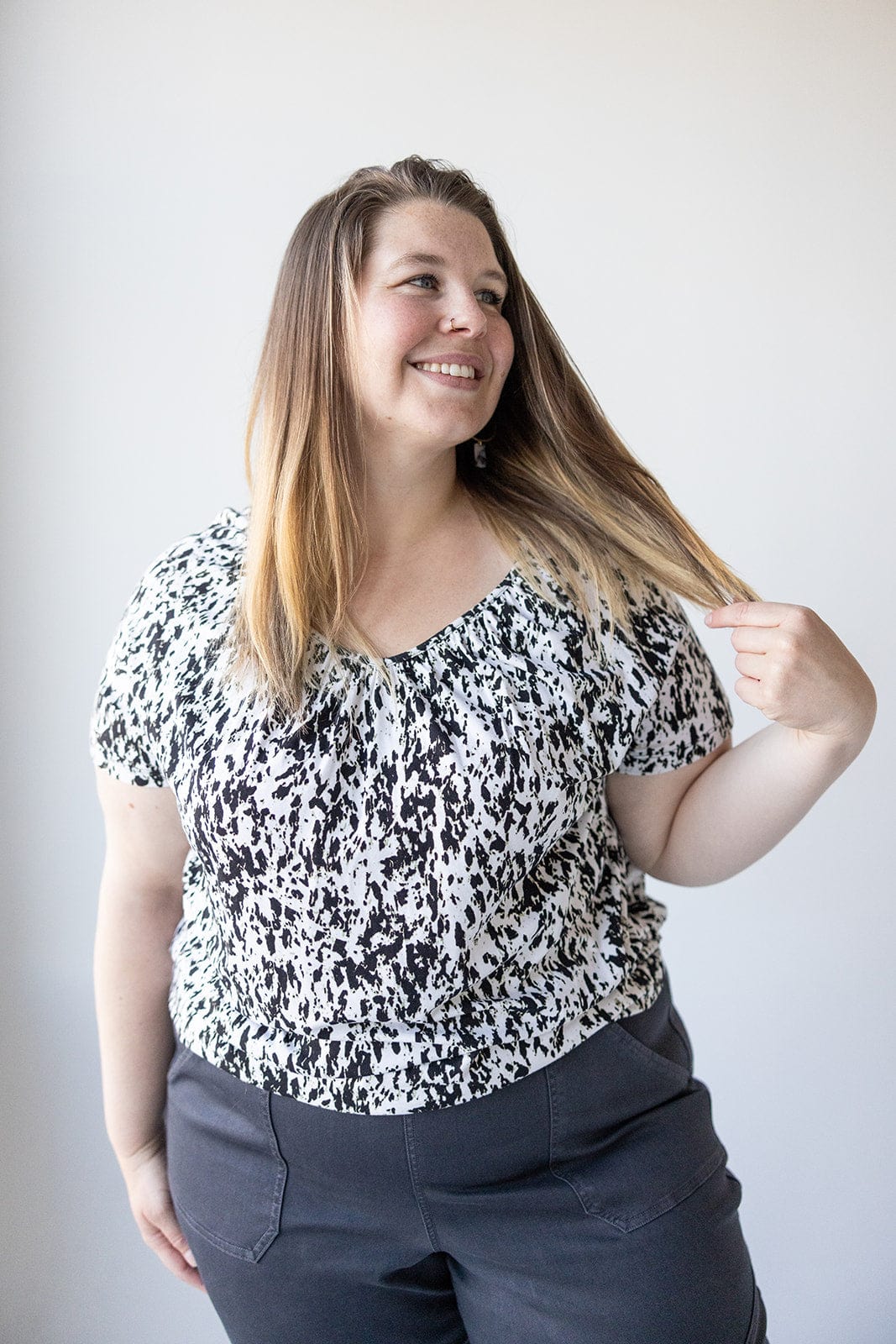 A person with long hair smiles and holds a strand of their hair. They are wearing a V-NECK TUNIC WITH ABSTRACT ANIMAL PRINT DETAIL by Love Marlow, paired perfectly with dark pants.