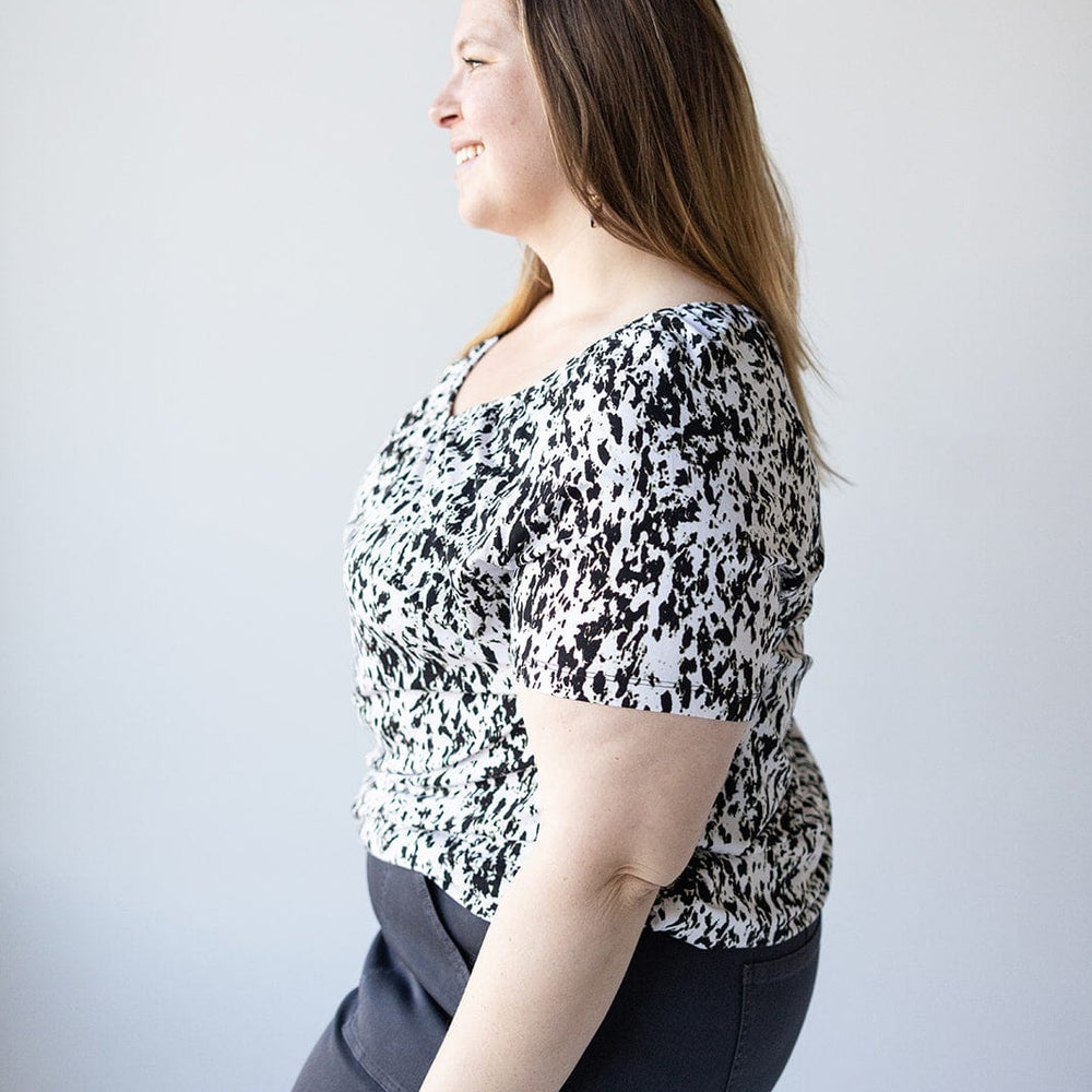 A woman with long brown hair, wearing a Love Marlow V-NECK TUNIC WITH ABSTRACT ANIMAL PRINT DETAIL and dark pants, is smiling while standing in profile against a white background.