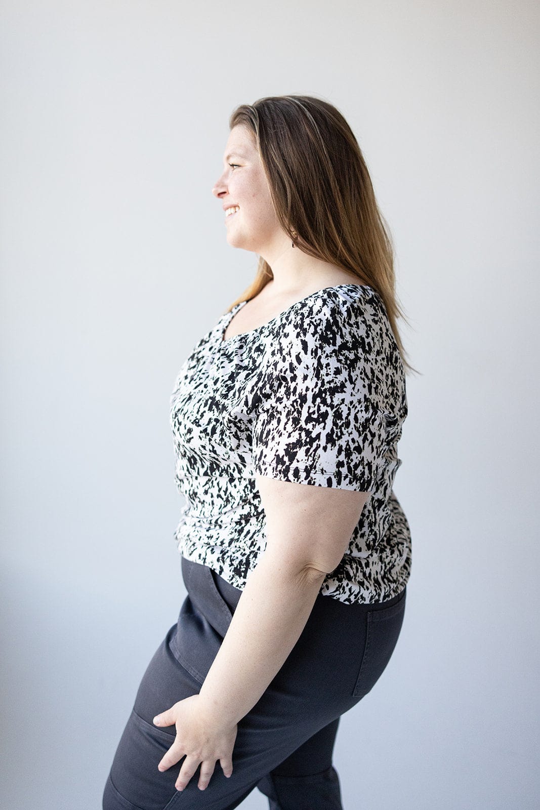 A woman with long brown hair, wearing a Love Marlow V-NECK TUNIC WITH ABSTRACT ANIMAL PRINT DETAIL and dark pants, is smiling while standing in profile against a white background.