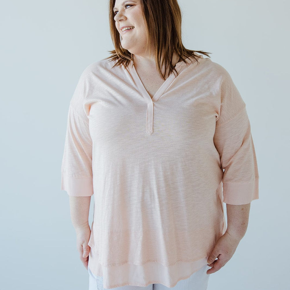 A person wearing a soft pink V-neck tunic with subtle hem detail and white pants stands against a plain background, looking to the side and smiling.