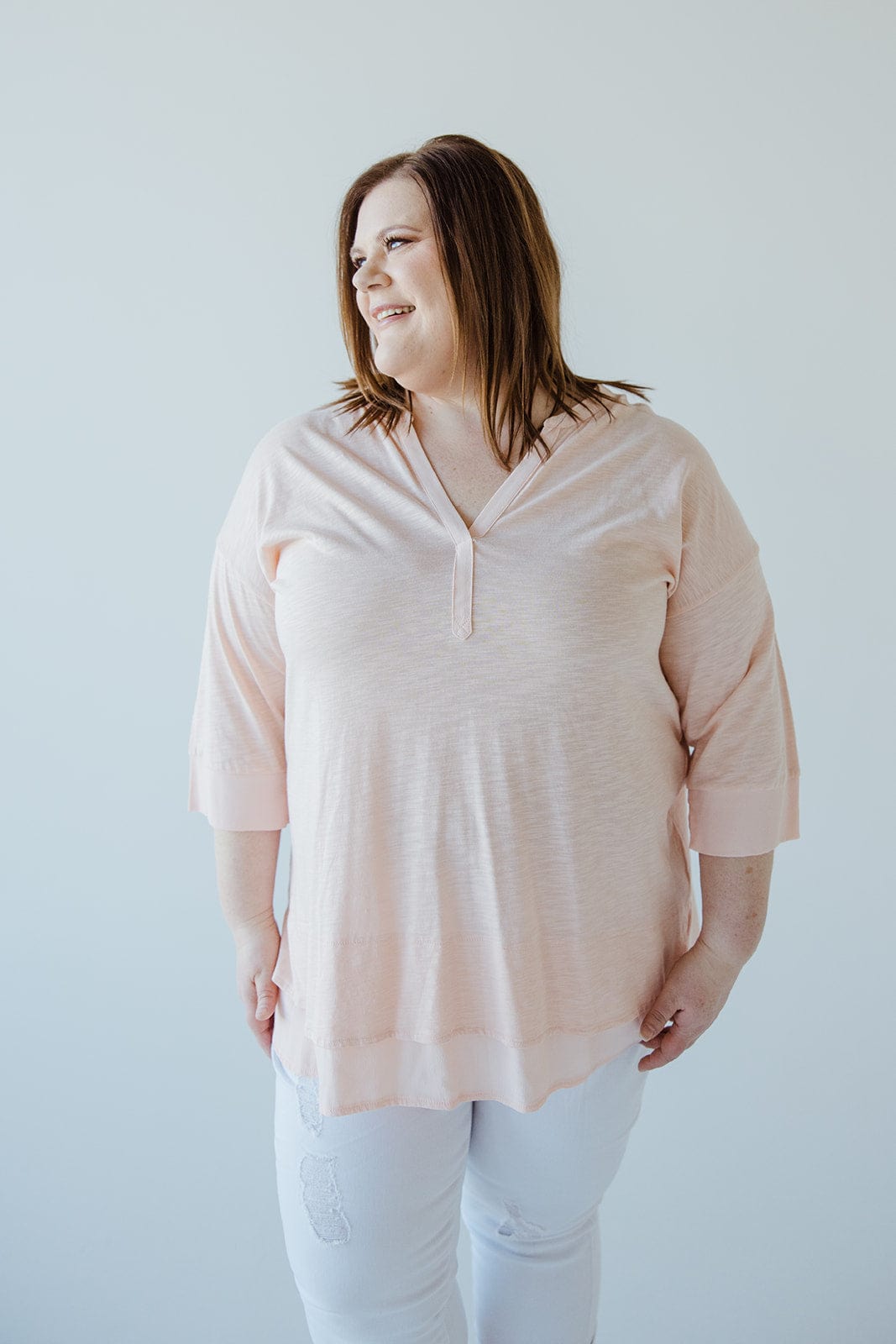 A person wearing a soft pink V-neck tunic with subtle hem detail and white pants stands against a plain background, looking to the side and smiling.