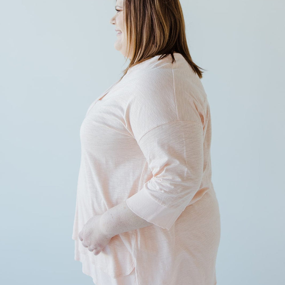 A person in a soft pink V-neck tunic and white pants, facing sideways against a plain white background, reveals subtle hem detail.