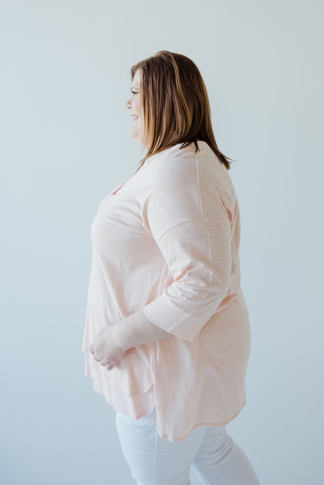 A person in a soft pink V-neck tunic and white pants, facing sideways against a plain white background, reveals subtle hem detail.