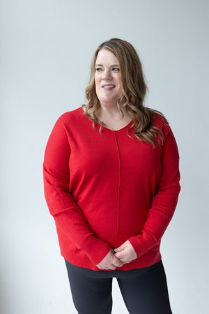 Individual with long hair wearing a Zenana V-Neck Front Seam Sweater in red, accompanied by black pants, stands against a white background, slightly smiling.