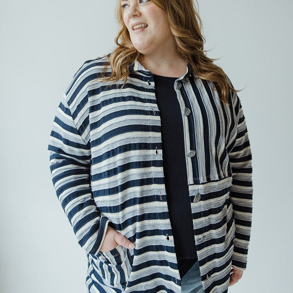 A woman wearing the VARYING STRIPE BUTTON-UP BLOUSE IN NAVY by neon buddha and jeans stands with a hand in her pocket, smiling and looking to her right.