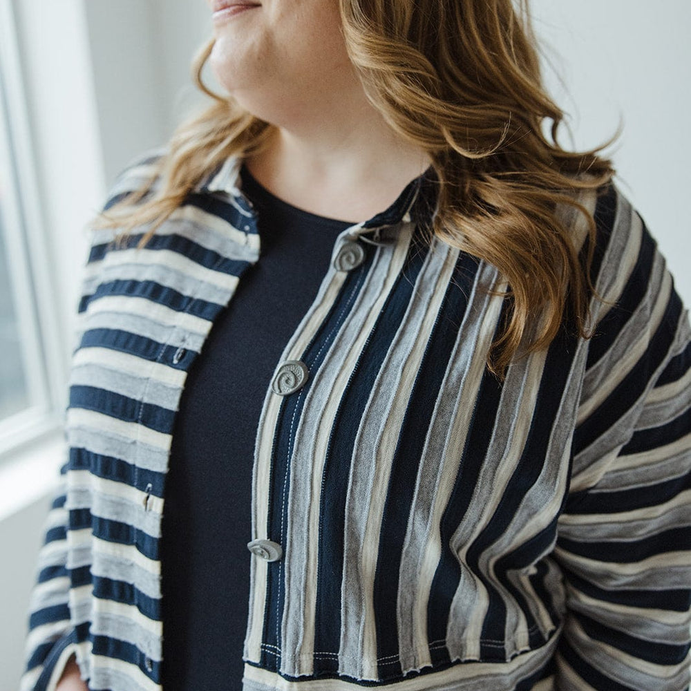 An individual with long hair wearing the VARYING STRIPE BUTTON-UP BLOUSE IN NAVY from neon buddha stands by a window, unique buttons softly glinting as they gaze outside.