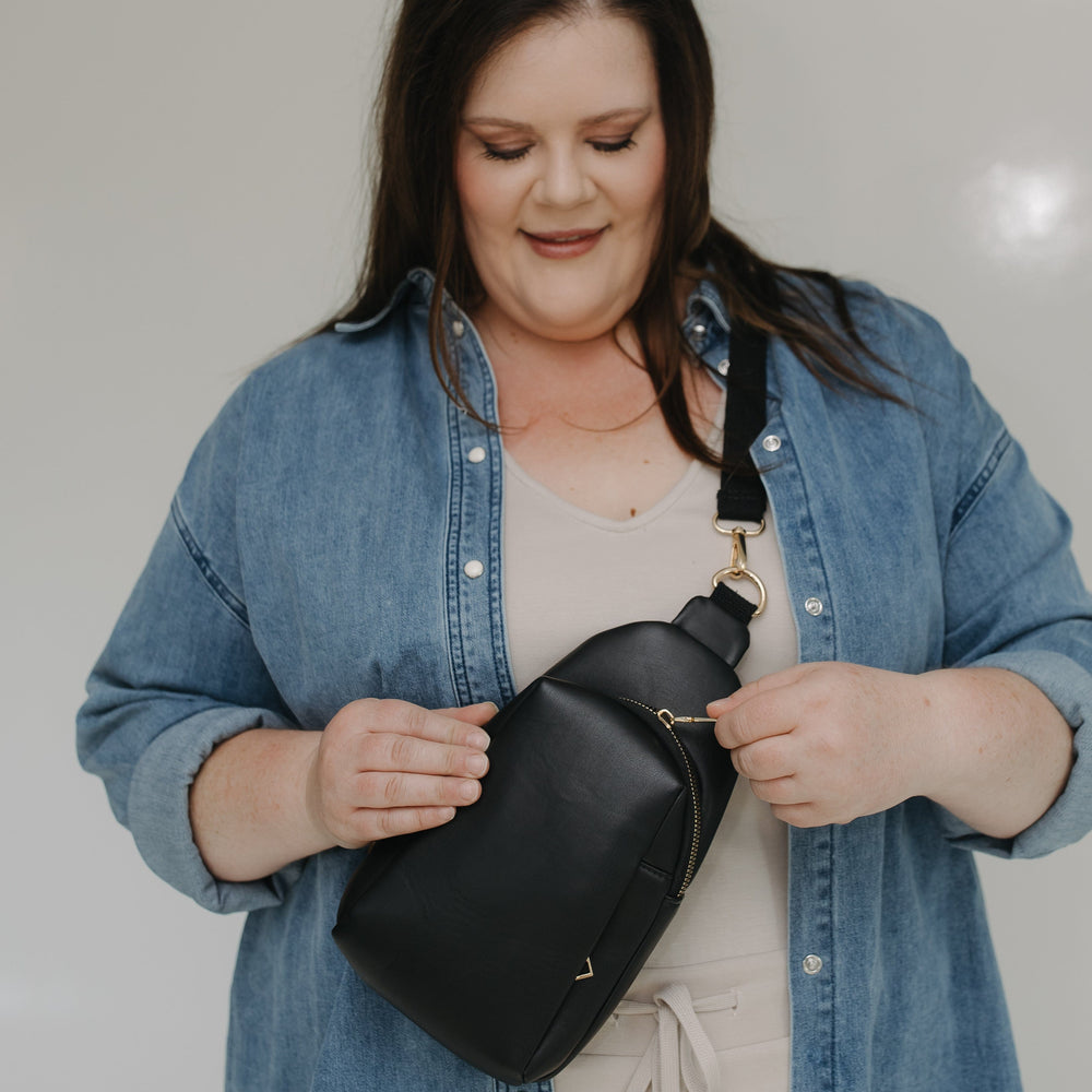 Woman wearing a denim jacket holding a sling bag against a plain background