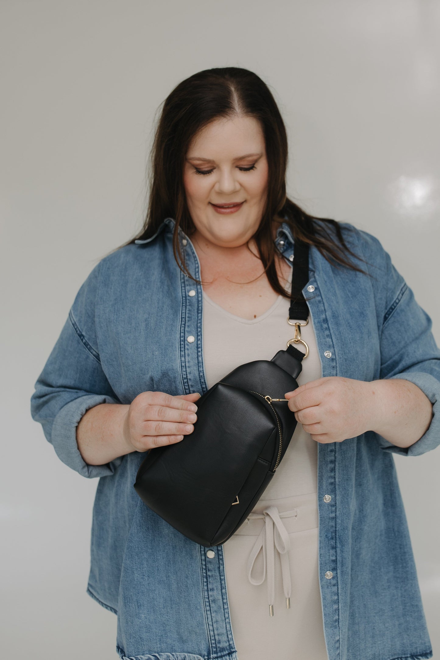 Woman wearing a denim jacket holding a sling bag against a plain background