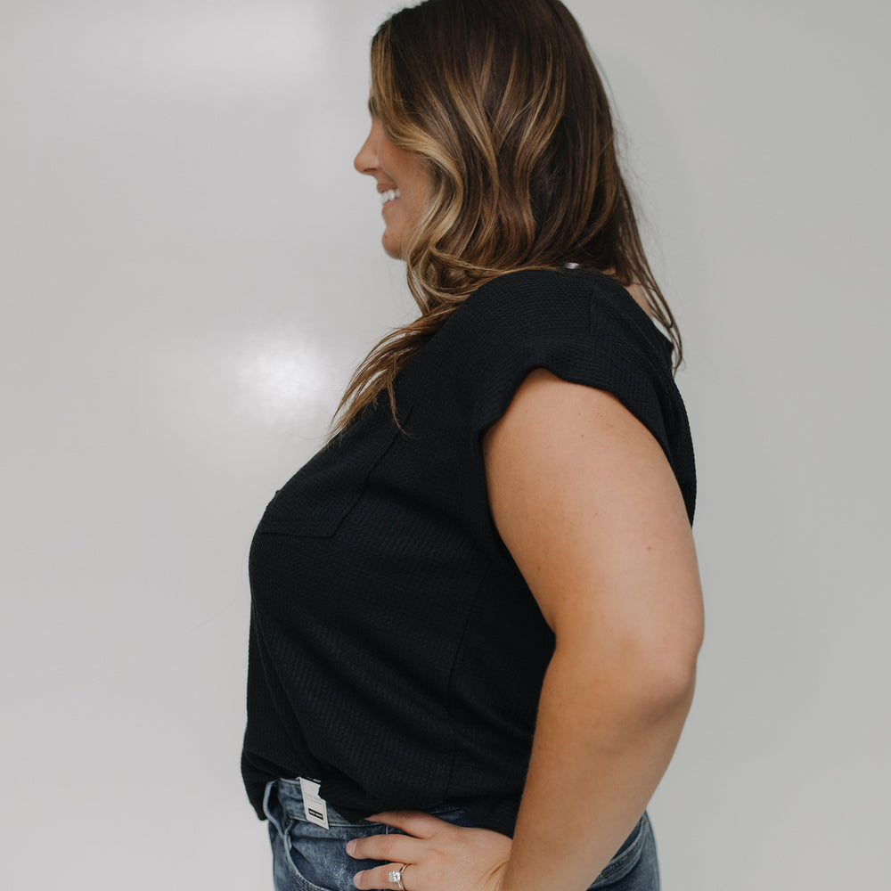 Woman wearing a black sleeveless top and blue jeans against a white wall.