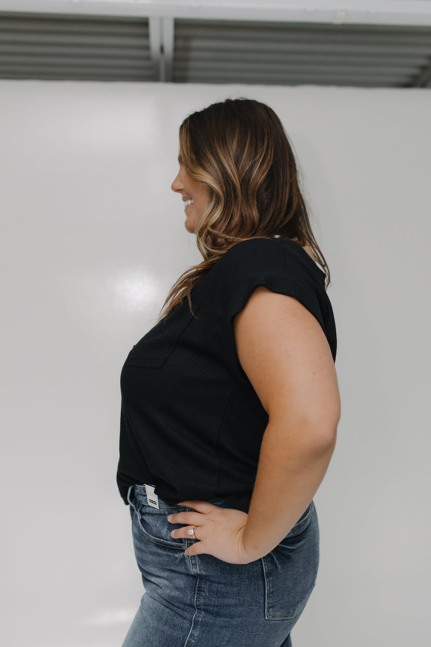 Woman wearing a black sleeveless top and blue jeans against a white wall.