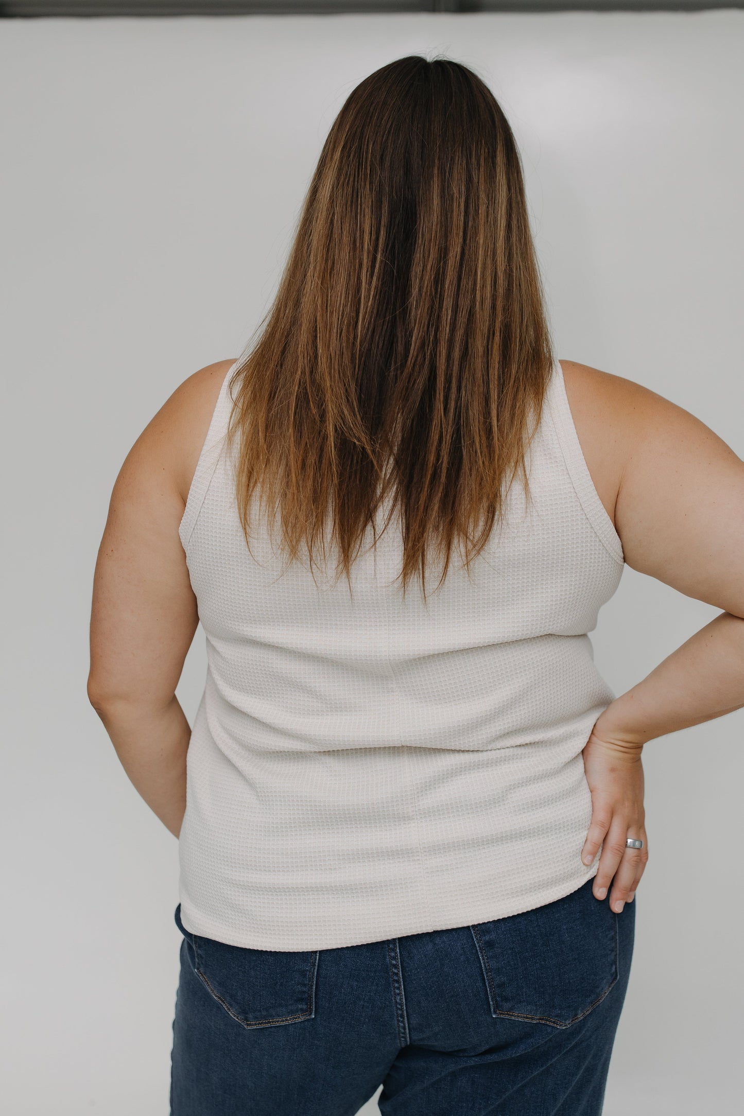 Person wearing a beige tank top and blue jeans against a plain background