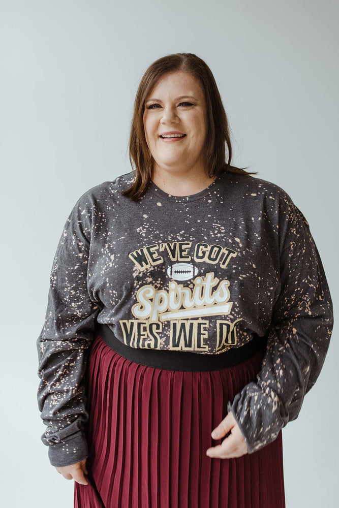 A person is smiling in a "WE'VE GOT SPIRITS YES WE DO TAILGATE" graphic tee by Love Marlow, paired with a maroon pleated skirt. The dark speckled design stands out against the plain background, capturing the essence of football season apparel made from ultra-soft material.