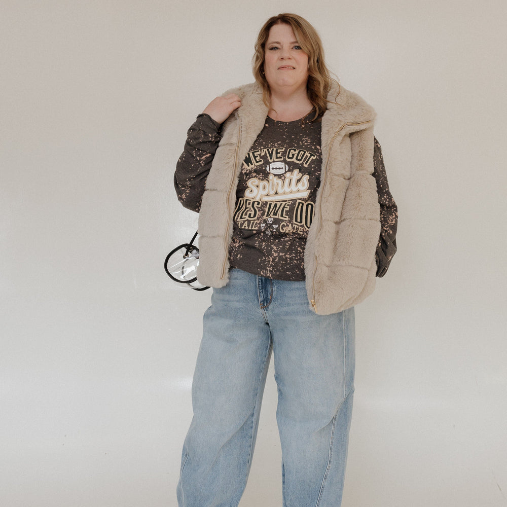 A person is smiling in a "WE'VE GOT SPIRITS YES WE DO TAILGATE" Love Marlow graphic tee by Love Marlow, paired with a maroon pleated skirt. The dark speckled design stands out against the plain background, capturing the essence of football season apparel made from ultra-soft material.