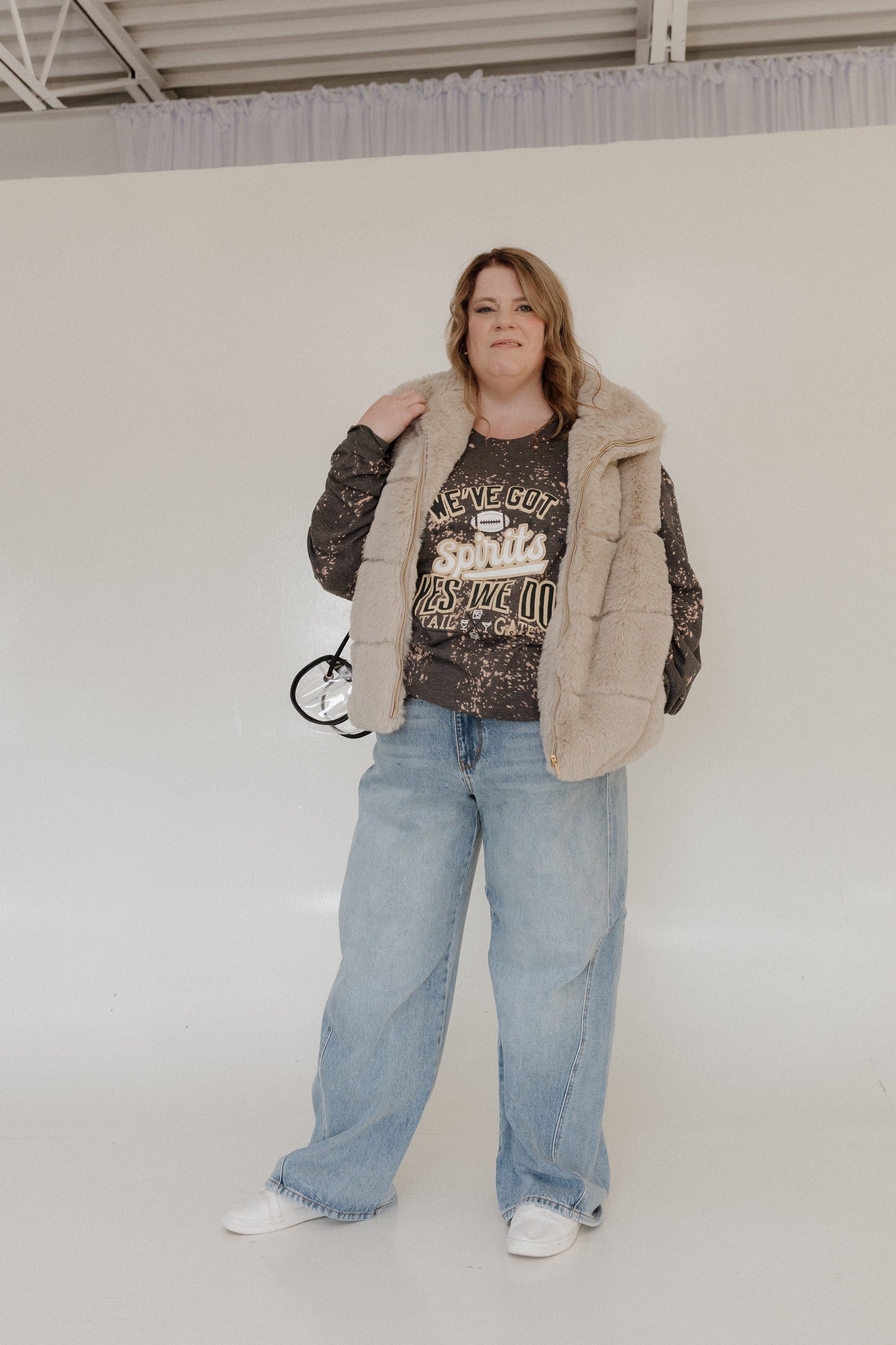 A person is smiling in a "WE'VE GOT SPIRITS YES WE DO TAILGATE" Love Marlow graphic tee by Love Marlow, paired with a maroon pleated skirt. The dark speckled design stands out against the plain background, capturing the essence of football season apparel made from ultra-soft material.