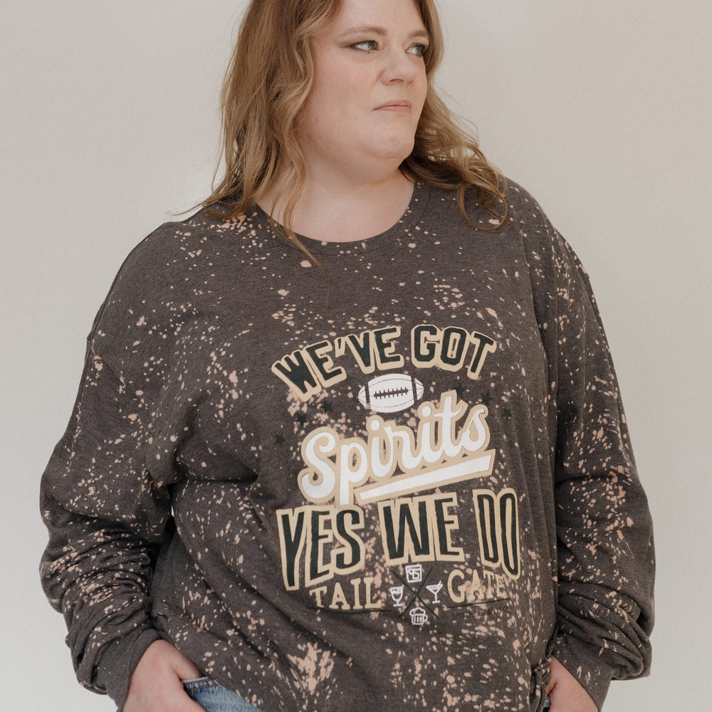 A person is smiling in a "WE'VE GOT SPIRITS YES WE DO TAILGATE" Love Marlow graphic tee by Love Marlow, paired with a maroon pleated skirt. The dark speckled design stands out against the plain background, capturing the essence of football season apparel made from ultra-soft material.