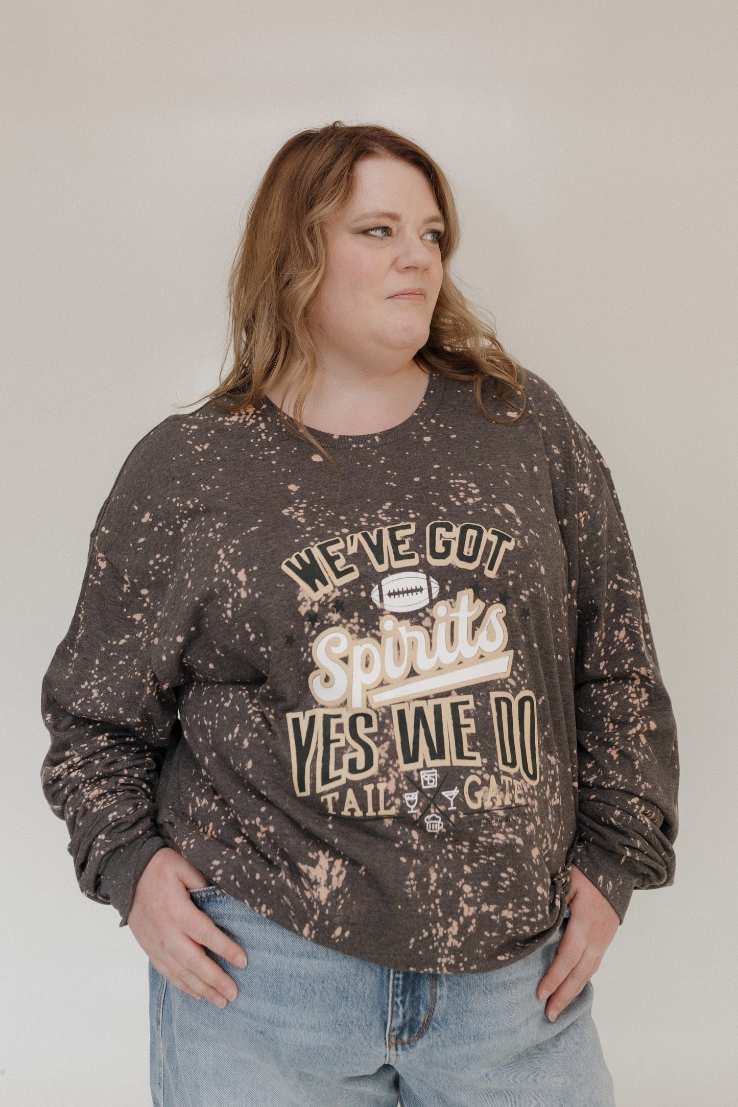 A person is smiling in a "WE'VE GOT SPIRITS YES WE DO TAILGATE" Love Marlow graphic tee by Love Marlow, paired with a maroon pleated skirt. The dark speckled design stands out against the plain background, capturing the essence of football season apparel made from ultra-soft material.