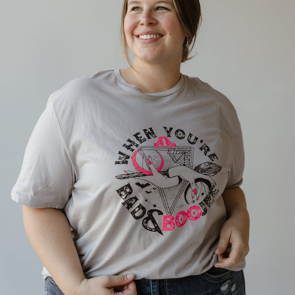 A person is smiling while wearing a graphic tee and jeans. The T-shirt, from Love Marlow, showcases a cowgirl hat, boots, and the phrase "WHEN YOU'RE BAD AND BOOJEE," making it perfect for capturing that bad and boojee vibe this Halloween season.
